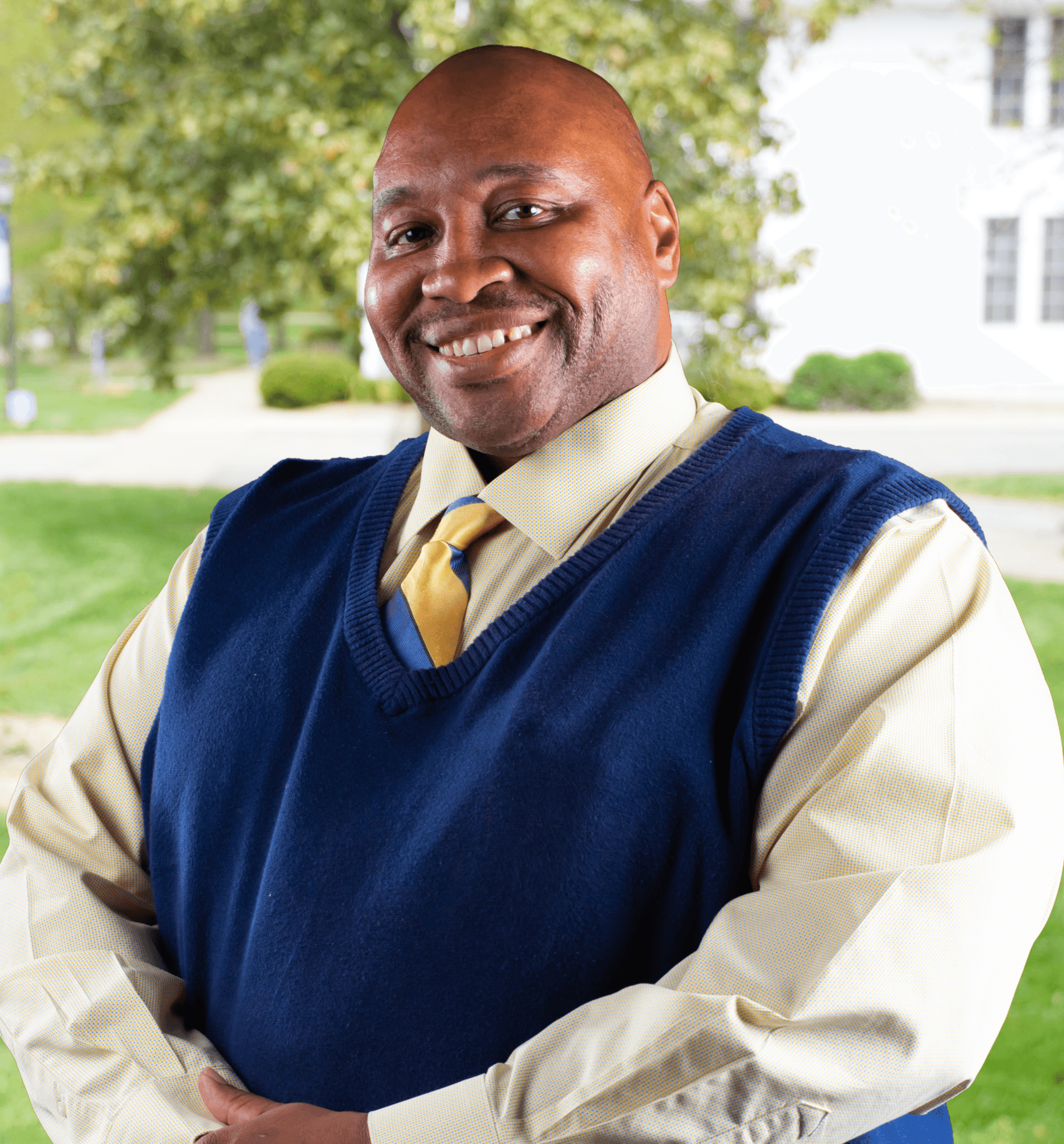 Portrait of Frank standing outdoors in front of Berea's admisisons building wearing a yellow button-down shirt, yellow tie, and navy sweater vest, with trees and a white building blurred in the background.